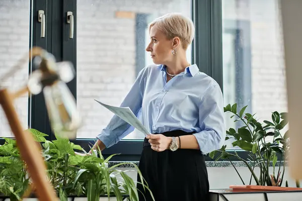 Middle-aged businesswoman with short hair standing by window, holding a piece of paper in office setting. — Stock Photo