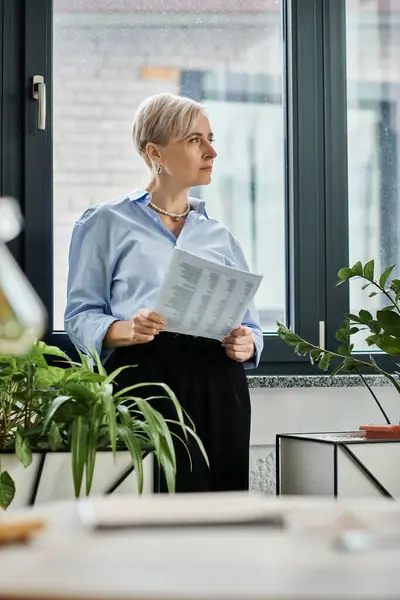 A middle-aged businesswoman with short hair reads a newspaper by a window in a tranquil office setting. — Stock Photo