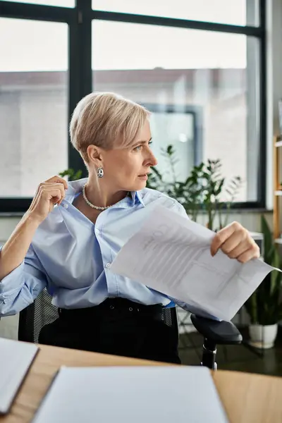 Una mujer de negocios de mediana edad con el pelo corto se sienta en un escritorio, sintiendo calor durante la menopausia - foto de stock