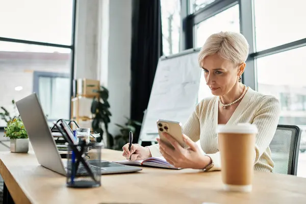 Una donna d'affari di mezza età con i capelli corti è seduto a un tavolo con un computer portatile e un telefono cellulare in un ambiente ufficio moderno. — Foto stock