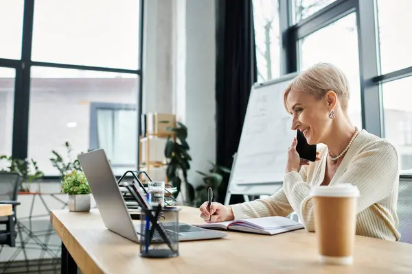 Middle-aged businesswoman with short hair multitasking by talking on phone and using laptop in modern office. — Stock Photo