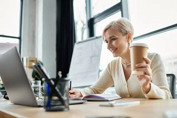 A middle aged businesswoman with short hair working diligently on her laptop while enjoying a cup of coffee at her desk. — Stock Photo