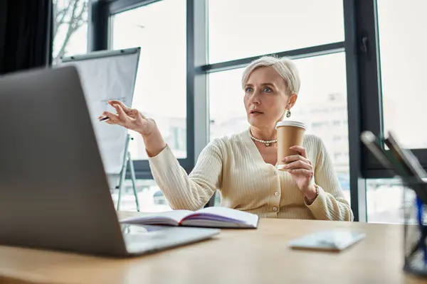 A middle-aged businesswoman with short hair, sitting at a desk, holding a cup of coffee in a modern office. — Stock Photo