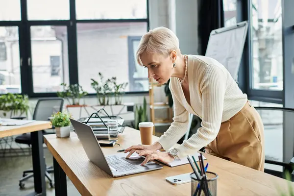 Middle-aged businesswoman with short hair focused on her laptop in a modern office setting. — Stock Photo