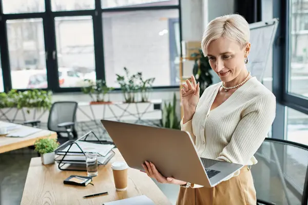 A middle-aged businesswoman with short hair stands in front of a laptop computer in a modern office setting. — Stock Photo
