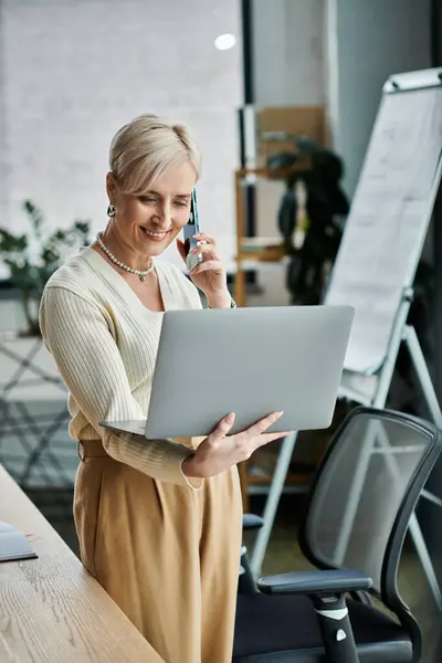 A middle-aged businesswoman with short hair simultaneously talks on a cell phone and works on a laptop in a modern office setting. — Stock Photo