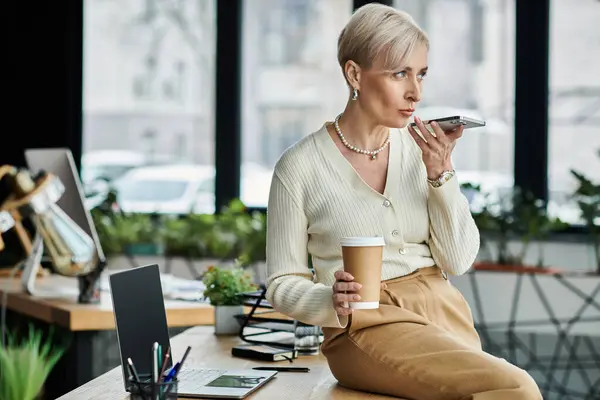 Middle-aged businesswoman with short hair sitting at a table, savoring a cup of coffee in a modern office setting. — Stock Photo