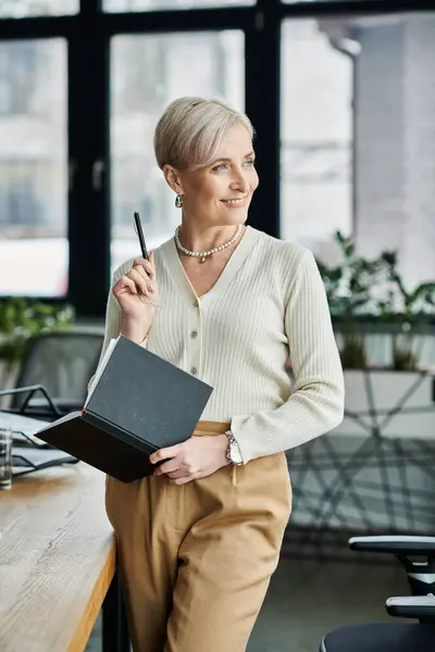 Middle-aged businesswoman with short hair holding a notebook and pen while standing in a modern office. — Stock Photo