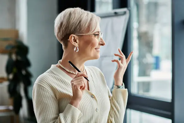 Empresária de meia-idade com cabelo curto, vestida com uma camisola branca, agita notas com uma caneta em um ambiente de escritório moderno. — Fotografia de Stock