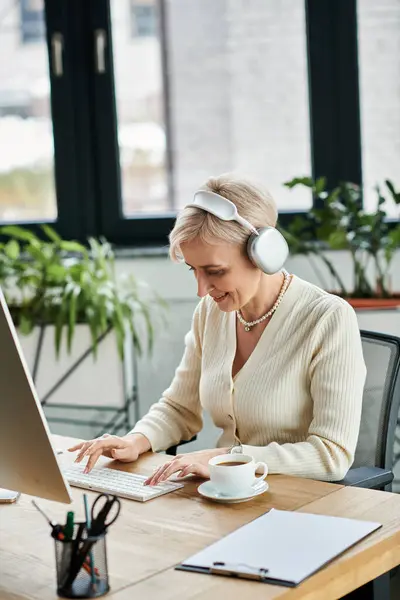 A middle-aged businesswoman with short hair engaged in work, wearing headphones at a modern office desk. — Stock Photo