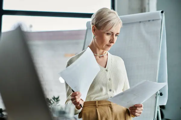 A middle-aged businesswoman with short hair holds a piece of paper in front of a computer screen. — Stock Photo