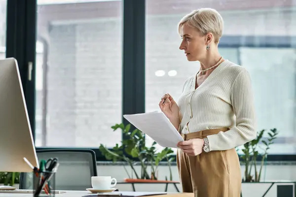 Middle-aged businesswoman with short hair stands in front of computer holding paper. — Stock Photo