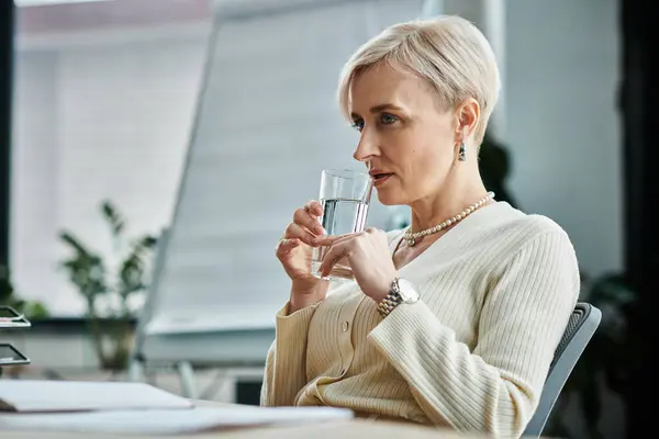 Middle aged businesswoman with short hair sitting at desk, peacefully holding a glass of water. — Stock Photo