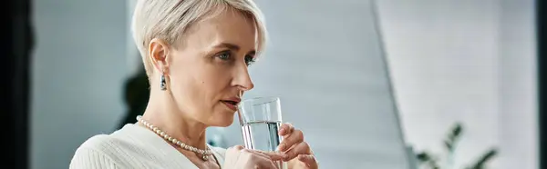 A middle aged businesswoman with short hair holds a glass of water in her hand in a modern office setting. — Stock Photo