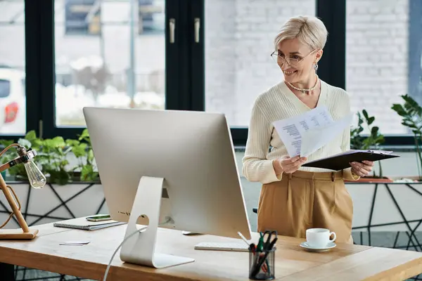 A middle aged businesswoman stands in front of a computer, holding a piece of paper while working in a modern office setting. — Stock Photo