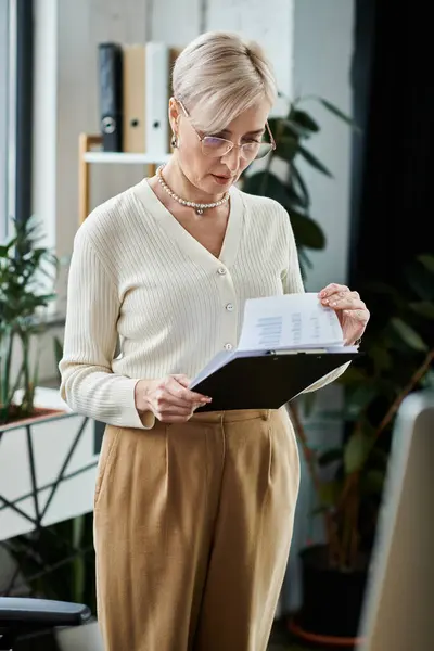 A middle aged businesswoman stands before a computer, holding a clipboard, immersed in work in a modern office setting. — Stock Photo