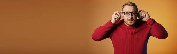 A dapper man in a red sweater gracefully holds a pair of glasses and headphones. — Stock Photo