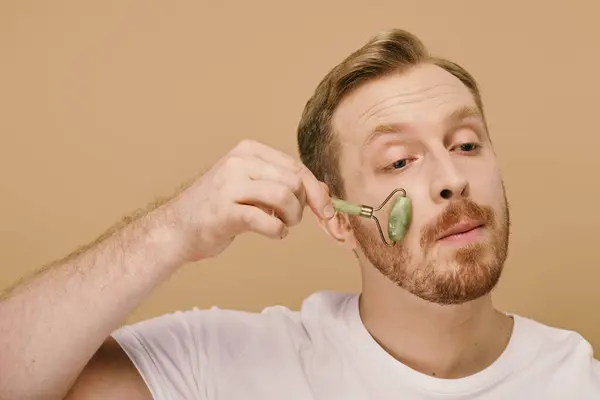 A man in casual attire gently using face roller for a skincare routine. — Stock Photo