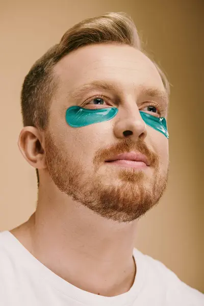 Un hombre con barba con parches de ojos azules se centra en su rutina de cuidado de la piel. - foto de stock