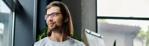 Schöner Kaukasier mit langen Haaren und Brille, der vor einem Fenster steht. — Stockfoto