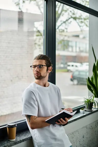Un apuesto hombre de negocios caucásico parado frente a una ventana, profundamente en pensamiento, sosteniendo un libro. - foto de stock