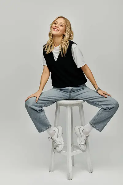 Young blonde woman sitting gracefully on white stool in stylish attire against grey background. — Stock Photo