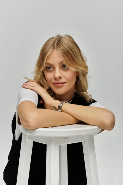 A stylish young woman with blonde hair sitting gracefully on a white stool against a plain grey backdrop. — Stock Photo