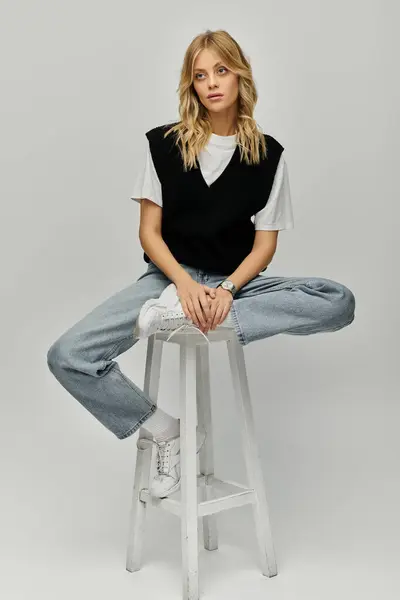 A stylish young woman with blonde hair sits elegantly atop a modern stool in casual attire against a solid grey backdrop. — Stock Photo