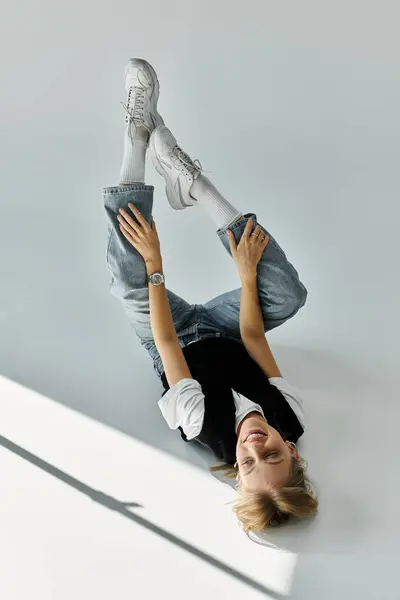 A young woman with blonde hair dressed in stylish casual attire lays gracefully on top of a white wall. — Stock Photo