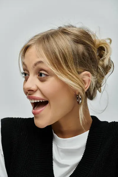 A young blonde woman smiles brightly while dressed in a black sweater on a grey background. — Stock Photo