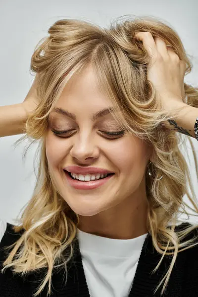 A young, beautiful woman smiling as she delicately holds her blonde hair in her hands against a grey background. — Stock Photo