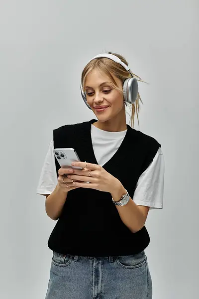 A young blonde woman in casual attire, wearing headphones, captivated by her cell phone. — Stock Photo