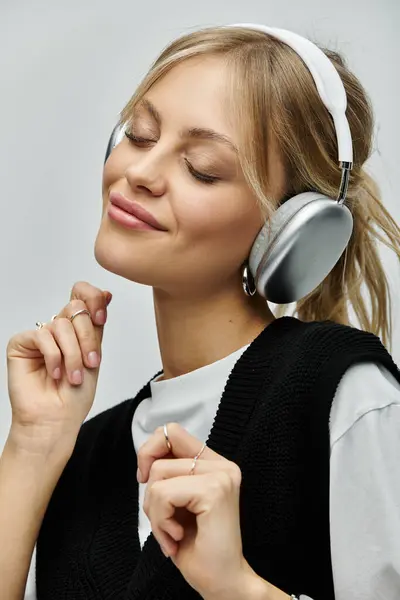 Una joven con estilo y cabello rubio con auriculares escucha música sobre un fondo gris. - foto de stock