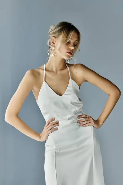Una bella giovane donna con i capelli biondi posa con grazia in un abito bianco su uno sfondo grigio. — Foto stock