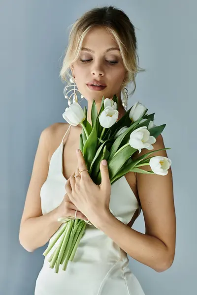A blonde woman in a white dress delicately holds a bouquet of flowers, exuding elegance and beauty. — Stock Photo