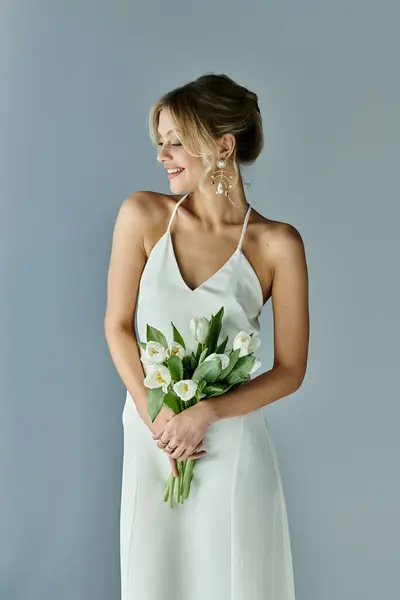 A beautiful, young woman with blonde hair holding a bouquet of flowers in a white dress on a grey background. — Stock Photo