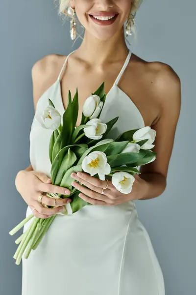 Beautiful blonde woman in elegant white dress standing with a bouquet of flowers in her hands on a grey background. — Stock Photo