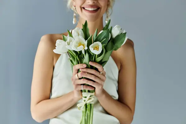 A blonde woman exudes elegance and beauty as she holds a bouquet of flowers in a white dress on a grey background. — Stock Photo