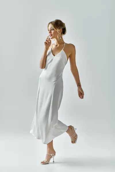 A young, beautiful blonde woman gracefully walks in a flowing white dress against a soft grey backdrop. — Stock Photo