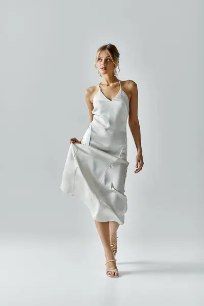 Una mujer joven y hermosa con el pelo rubio camina elegantemente en un vestido blanco que fluye contra un telón de fondo neutral. - foto de stock