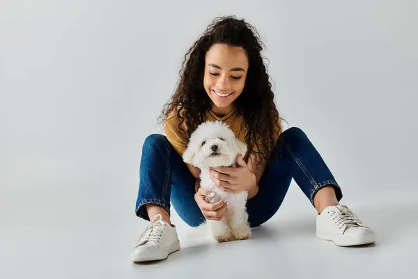 A young woman sits calmly on the floor, cuddling a fluffy white Bichon Frise. — Stock Photo