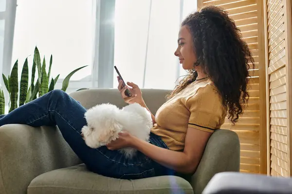 A woman sitting on a couch, cuddling her dog and checking her phone. — Stock Photo