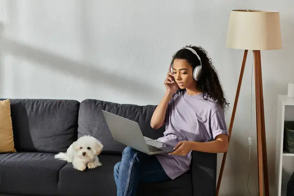 A woman absorbed in her laptop while seated on a couch, accompanied by her bichon frise dog. — Stock Photo