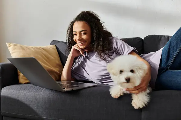 A young woman sits on a couch with a laptop, accompanied by her bichon frise dog. — Stock Photo