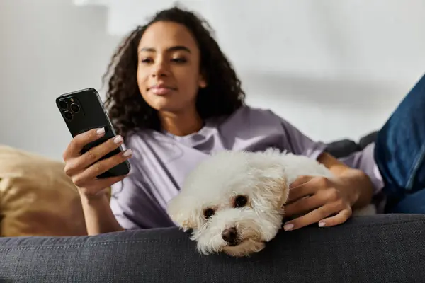 A woman relaxes on a couch, holding her bichon frise dog and a cell phone. — Stock Photo