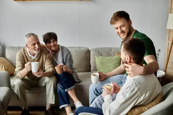 A gay couple sits on a sofa, embraced and holding cups, with parents in the background. — стоковое фото