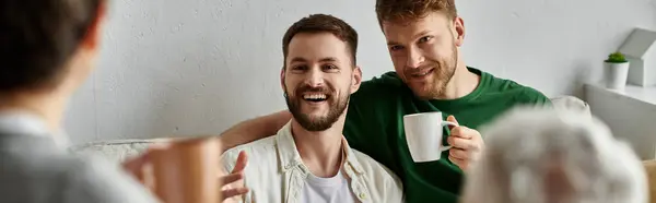 A gay couple smiles and holds mugs while sitting on a couch in their home, likely meeting with family. — Stock Photo