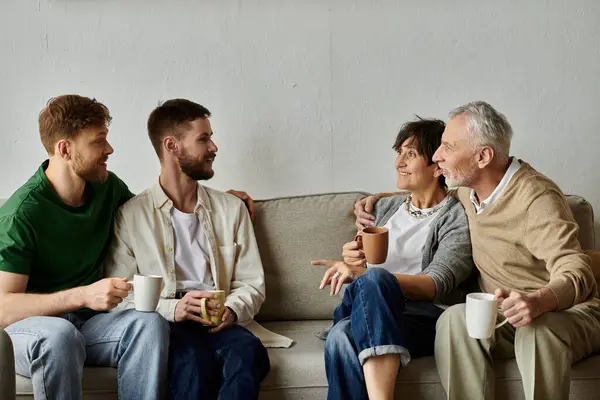 A gay couple sits on a couch with parents, all enjoying coffee together. — стоковое фото