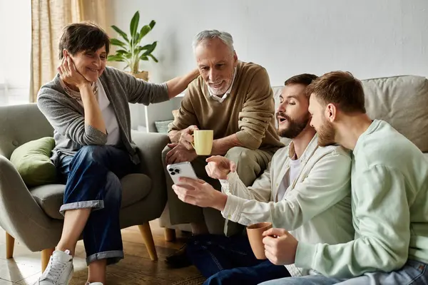 Um casal gay se senta em um sofá com os pais em sua casa, compartilhando notícias ou vendo algo em um telefone. — Fotografia de Stock