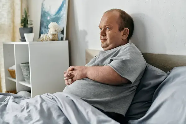 A man with inclusivity sits in bed, lost in thought, as the morning light filters through the window. — Stock Photo